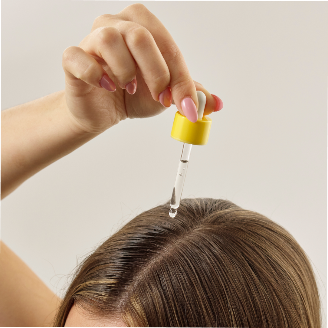 A hand holding a dropper from Growth Bomb USs Hair Growth Support Bundle applies serum to a parted brown-haired scalp. Nails painted pink stand out against the plain light background.