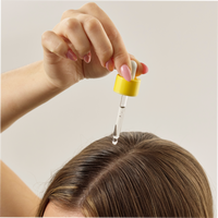 A hand holding a dropper from Growth Bomb USs Hair Growth Support Bundle applies serum to a parted brown-haired scalp. Nails painted pink stand out against the plain light background.