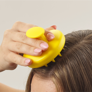 A person enjoys hair care with a Growth Bomb Silicone Scalp Brush in yellow, showcasing short spikes. Their long brown hair flows elegantly, while their soft pink manicured nails stand out against a neutral backdrop.