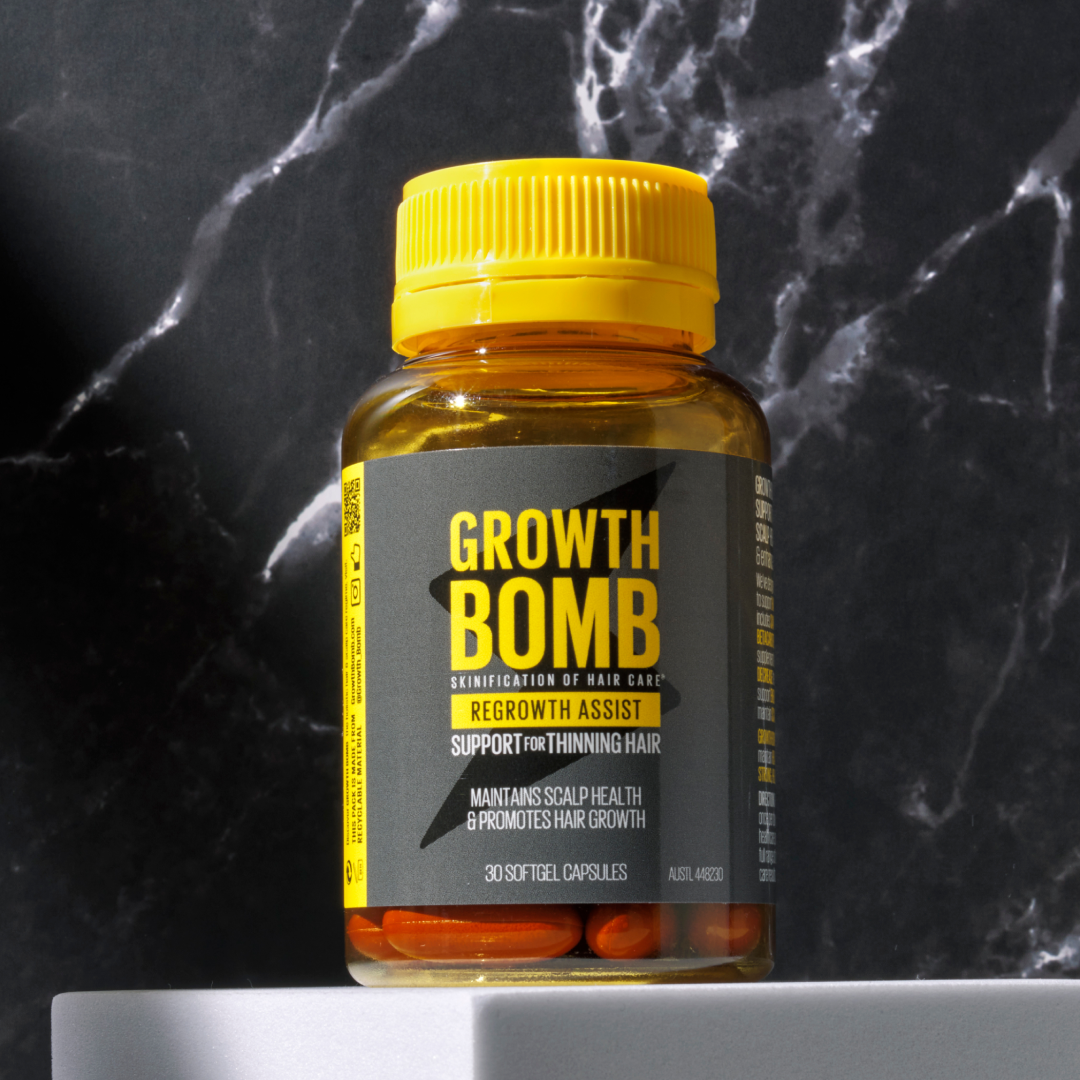 A bottle of Growth Bomb USs Regrowth Assist Support for Thinning Hair Supplement sits on a marble surface, featuring benefits for hair thickness and scalp health. The amber bottle with a yellow lid contains 30 softgel capsules designed to boost hair growth and vitality.