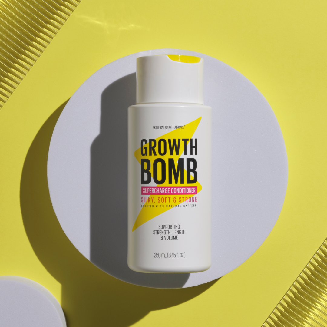 A bottle from Growth Bomb USs Hair Growth Support Bundle, infused with growth vitamins and caffeine for silky, strong hair, sits on a round white platform. It boasts a sleek white design, bold black text on a bright yellow background.