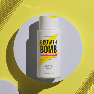 A bottle from Growth Bomb USs Hair Growth Support Bundle, infused with growth vitamins and caffeine for silky, strong hair, sits on a round white platform. It boasts a sleek white design, bold black text on a bright yellow background.
