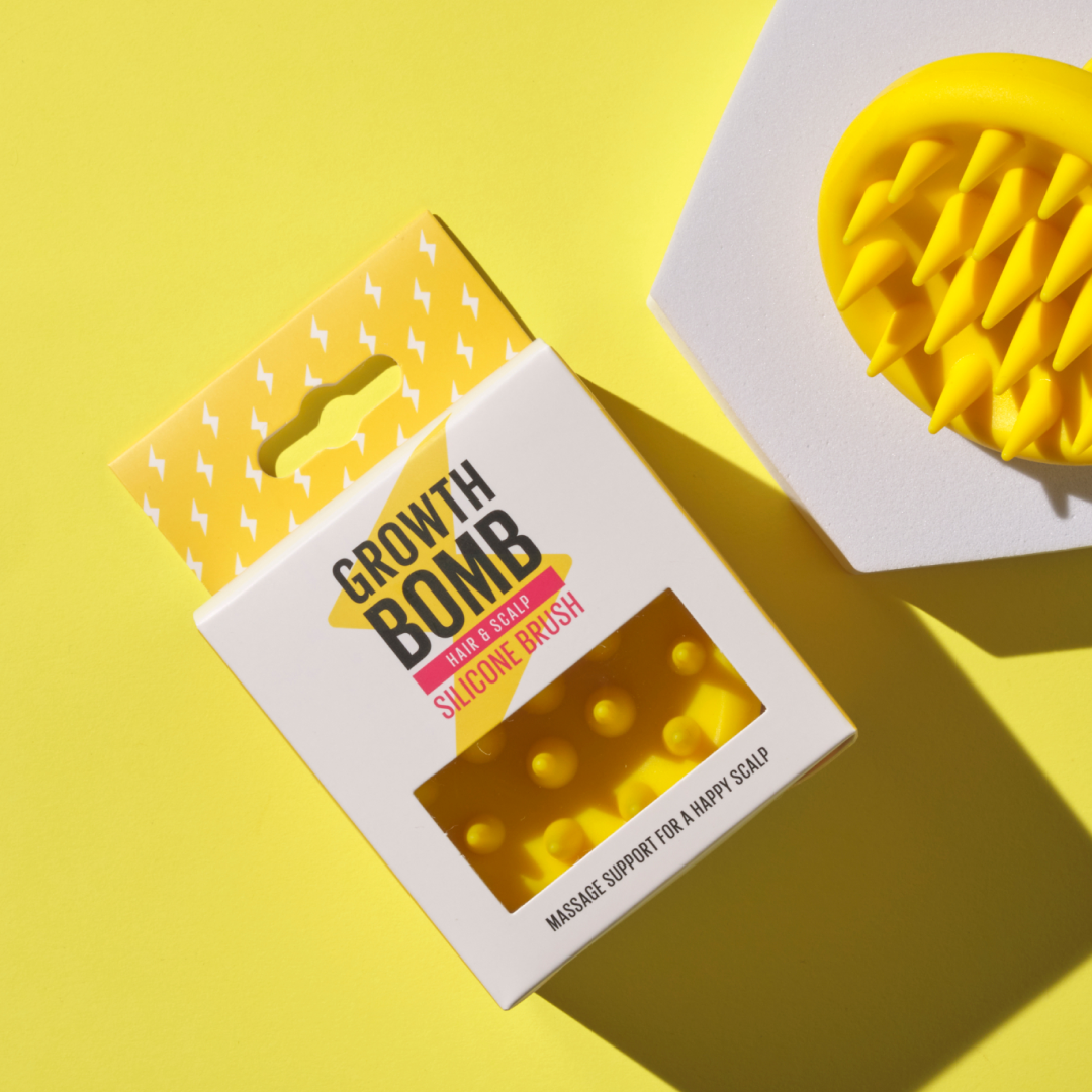 A yellow silicone scalp brush with spikes, partially shown beside its packaging on a vibrant yellow backdrop, displays Growth Bomb Silicone Scalp Brush with small white lightning bolts, emphasizing its role in enhancing hair and scalp care while aiding exfoliation.