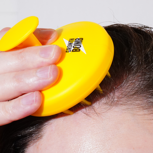 A hand uses a yellow Growth Bomb Silicone Scalp Brush on wet hair, showcasing the logo. This brush, ideal for exfoliating the scalp, is held firmly by fingers as it enhances hair and scalp care.