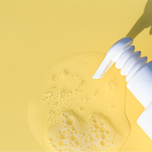 A white Smoothing Support Bundle bottle from Growth Bomb US has a flip cap that dispenses Yerba Maté-infused liquid soap onto a yellow surface, creating a bubbly puddle. The bottles shadow is visible behind the cap.