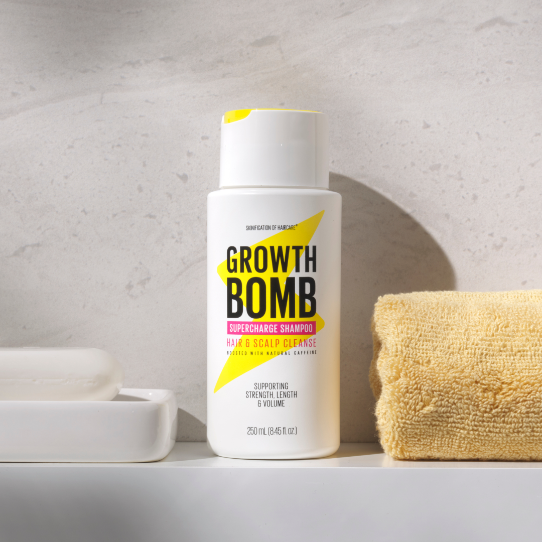 A white bottle of Growth Bomb USs Hair Growth Support Bundle, displaying bright yellow and pink text, sits on a bathroom counter next to a yellow towel, casting shadows on the light wall. Enhance your hair growth journey by adding our Hair Growth Vitamins for optimal results.