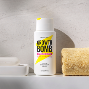 A white bottle of Growth Bomb USs Hair Growth Support Bundle, displaying bright yellow and pink text, sits on a bathroom counter next to a yellow towel, casting shadows on the light wall. Enhance your hair growth journey by adding our Hair Growth Vitamins for optimal results.