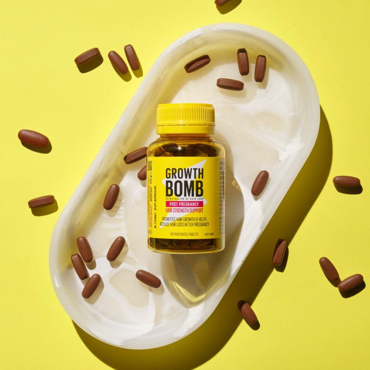 A yellow bottle labeled Post Pregnancy Hair Strength Support Supplement by Growth Bomb US sits on a white dish against a vibrant yellow background, highlighting its role in fighting hair loss. Brown tablets are scattered nearby, promising improved hair growth and strength.